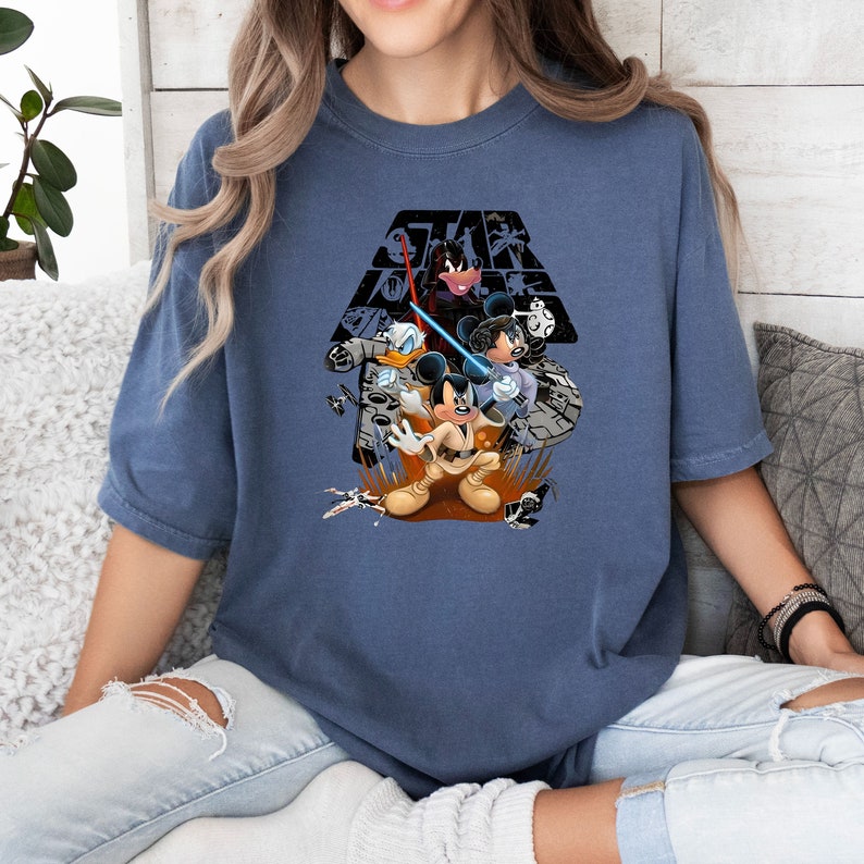 May include: A blue t-shirt with a graphic of Mickey Mouse, Donald Duck, and Goofy dressed as Star Wars characters. The graphic includes the Star Wars logo and a background of starships.