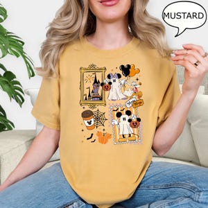 Może przedstawiać: Musztardowa koszulka z grafiką o tematyce Halloween. Grafika zawiera wizerunki Myszki Miki i Myszki Minnie przebranych za duchy, nawiedzony zamek, filiżankę kawy oraz napis "Always Magic".