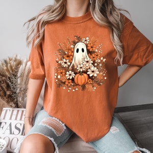 Op de afbeelding: Een oranje T-shirt met een witte geest en een pompoen omringd door oranje en witte bloemen.