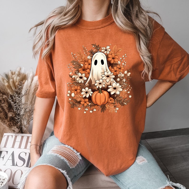 Op de afbeelding: Een oranje T-shirt met een witte geest en een pompoen omringd door oranje en witte bloemen.