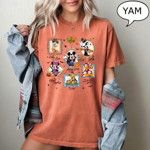 Puede incluir: Camiseta naranja con retratos enmarcados de personajes de Disney con tem&aacute;tica de Halloween. Mickey Mouse, Minnie Mouse, Donald Duck, Goofy, Pluto y Daisy Duck est&aacute;n representados con disfraces. La camiseta tiene la palabra "YAM" en una burbuja.