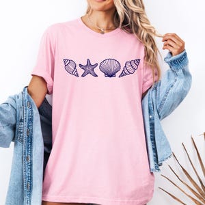 May include: A pink t-shirt with a navy blue outline of a seashell, starfish, and two more seashells.