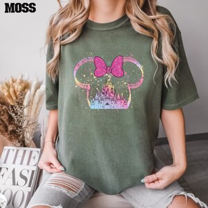 Puede incluir: Una camiseta verde con un lazo rosa y un gr&aacute;fico de castillo blanco. El castillo est&aacute; rodeado de un patr&oacute;n de salpicaduras de color arco&iacute;ris.