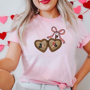 May include: Pink t-shirt with a heart-shaped locket featuring Mickey and Minnie Mouse. The locket is attached to a pink ribbon with a bow.