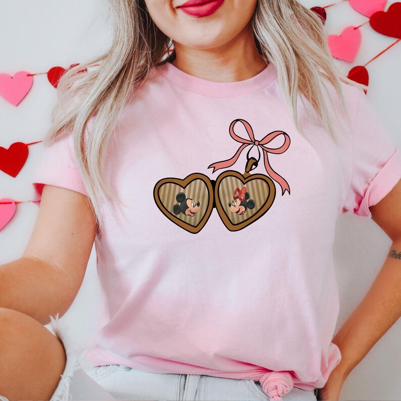 May include: Pink t-shirt with a heart-shaped locket featuring Mickey and Minnie Mouse. The locket is attached to a pink ribbon with a bow.