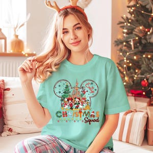 May include: A light teal t-shirt featuring a Disney-themed Christmas design. The design includes Mickey Mouse ears, a castle, and various Disney characters. The text "Christmas Squad" is displayed below the characters, with the name "Emily" above the characters.