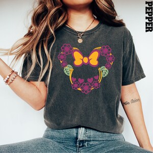 May include: A dark gray t-shirt with a floral design featuring a pink bow and yellow accents. The design is reminiscent of a popular cartoon character's head.