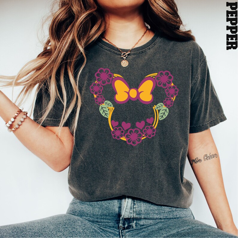 May include: A dark gray t-shirt with a floral design featuring a pink bow and yellow accents. The design is reminiscent of a popular cartoon character's head.