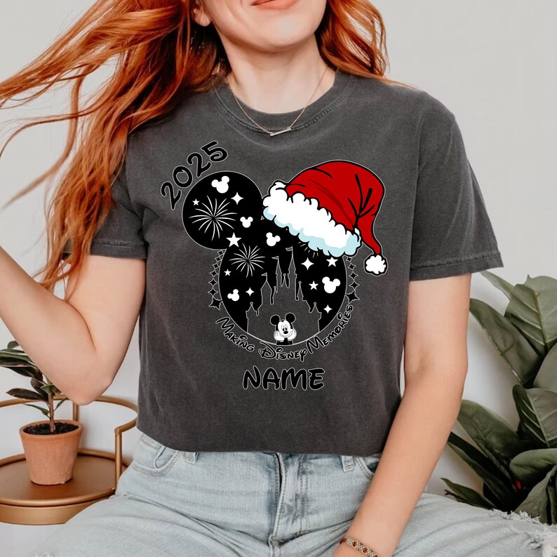 Peut inclure: Un t-shirt noir avec un motif de t&ecirc;te de Mickey Mouse. La t&ecirc;te de Mickey Mouse porte un chapeau de P&egrave;re No&euml;l et a des feux d'artifice et un ch&acirc;teau &agrave; l'int&eacute;rieur. Le texte "2025 Making Disney Memories NAME" est imprim&eacute; sur le t-shirt.
