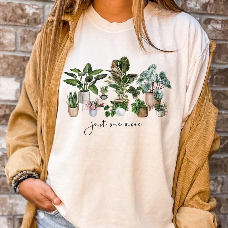 May include: A white t-shirt with a watercolor illustration of potted plants and the text "just one more".