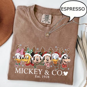 Puede incluir: Camiseta color espresso con un gr&aacute;fico festivo de personajes de Disney con cuernos de reno y bufandas. La camiseta incluye el texto "Mickey & Co. Est. 1928". Un collar plateado est&aacute; drapeado sobre la camiseta.