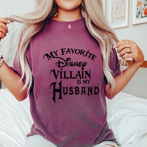 Pu&ograve; includere: Una maglietta viola con il testo "My Favorite Disney Villain Is My Husband" in lettere nere.