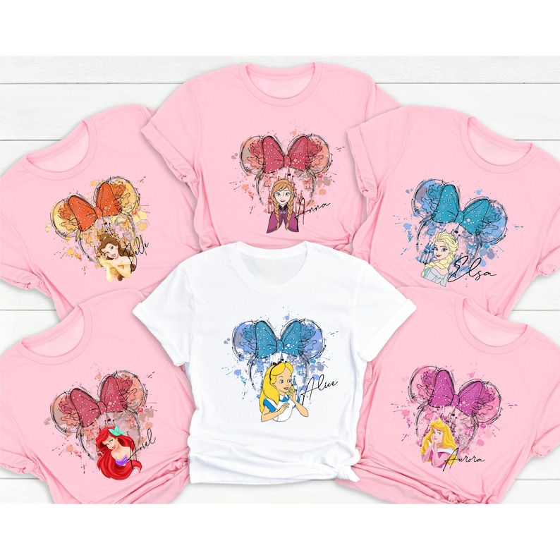 Peut inclure: Six t-shirts roses et blanches avec un motif aquarelle de princesses Disney avec des oreilles de souris. Chaque t-shirt a une princesse diff&eacute;rente et le nom "Anna", "Elsa", "Ariel", "Belle", "Alice" et "Aurora" est &eacute;crit sur chaque t-shirt.