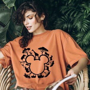 Puede incluir: Una camiseta naranja quemada con una silueta negra de la cabeza de Mickey Mouse. La silueta est&aacute; formada por varios animales, entre ellos una jirafa, una cebra y un elefante, todos rodeados de &aacute;rboles.