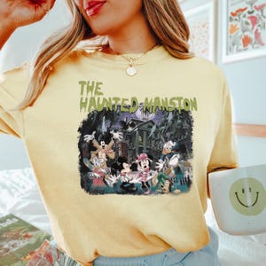 May include: A yellow t-shirt with a graphic print of Mickey Mouse and friends in front of the Haunted Mansion. The text "The Haunted Mansion" is printed in green on the shirt.