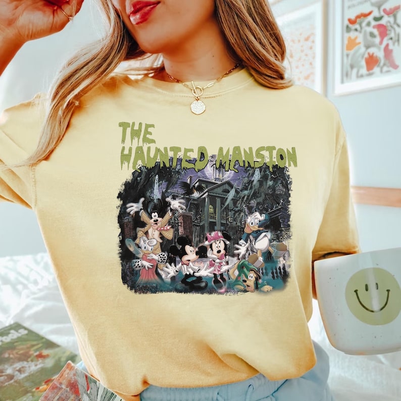 May include: A yellow t-shirt with a graphic print of Mickey Mouse and friends in front of the Haunted Mansion. The text "The Haunted Mansion" is printed in green on the shirt.