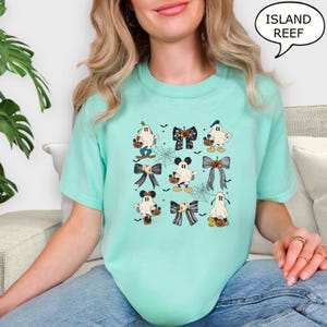 Puede incluir: Camiseta de color verde azulado claro con una cuadr&iacute;cula de gr&aacute;ficos tem&aacute;ticos de Halloween. Los gr&aacute;ficos incluyen fantasmas de dibujos animados, telara&ntilde;as y lazos con dise&ntilde;os de calabazas y Mickey Mouse. El texto "ISLAND REEF" est&aacute; en una burbuja.