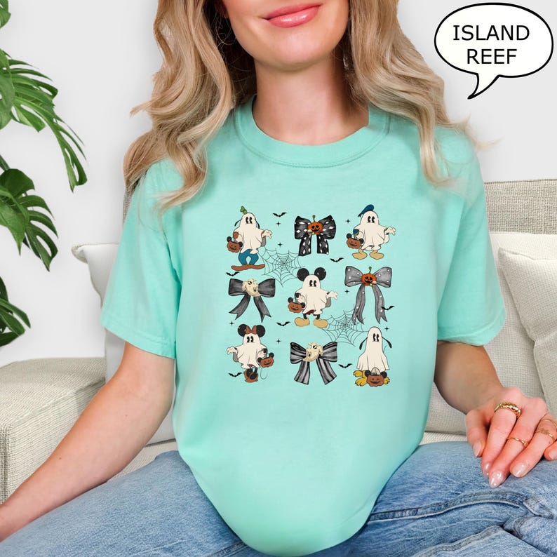 Puede incluir: Camiseta de color verde azulado claro con una cuadr&iacute;cula de gr&aacute;ficos tem&aacute;ticos de Halloween. Los gr&aacute;ficos incluyen fantasmas de dibujos animados, telara&ntilde;as y lazos con dise&ntilde;os de calabazas y Mickey Mouse. El texto "ISLAND REEF" est&aacute; en una burbuja.