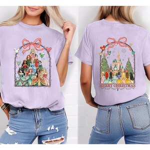 May include: A lilac t-shirt with a graphic of Disney princesses within a decorative arch and bow. The back displays a castle, Christmas trees, and the words "MERRY CHRISTMAS". The shirt is a comfortable, casual style.