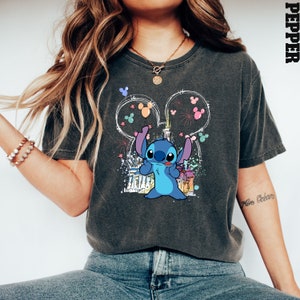 May include: A dark grey t-shirt with a blue Stitch character graphic. Stitch is standing in front of a white outline of the Cinderella Castle with fireworks and Mickey Mouse ears. The text "More Beers" is written on the right sleeve of the shirt.