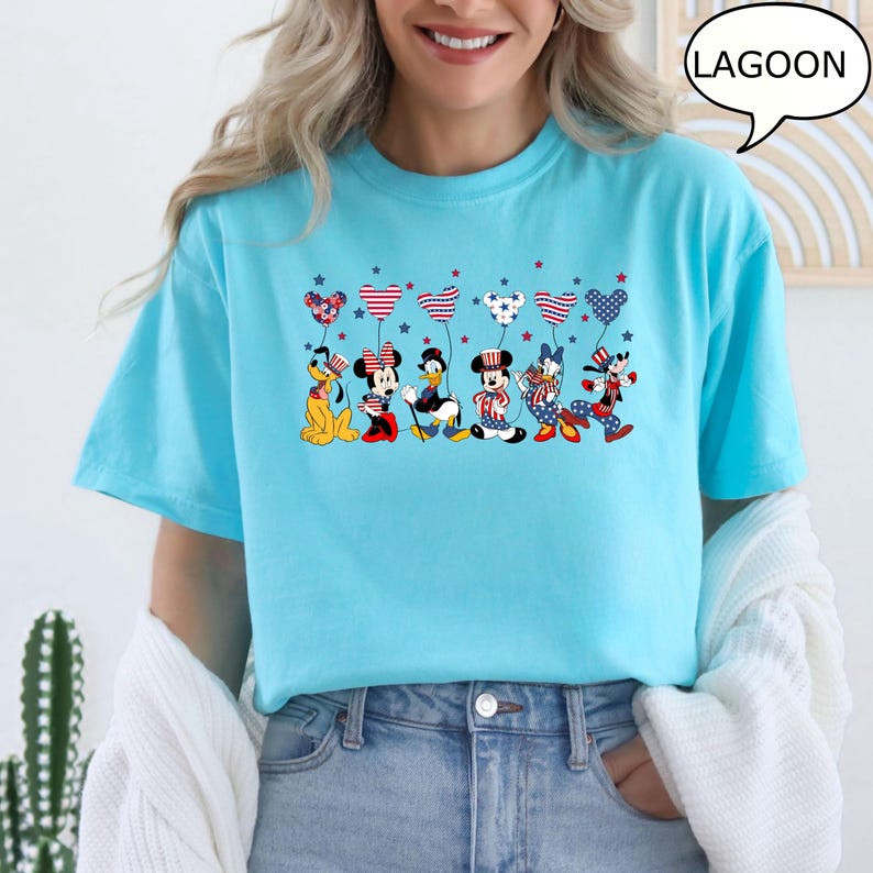 Puede incluir: Camiseta azul claro con personajes de dibujos animados sosteniendo globos en forma de coraz&oacute;n con dise&ntilde;os rojos, blancos y azules. La palabra "LAGOON" est&aacute; en una burbuja. Manga corta y corte holgado.