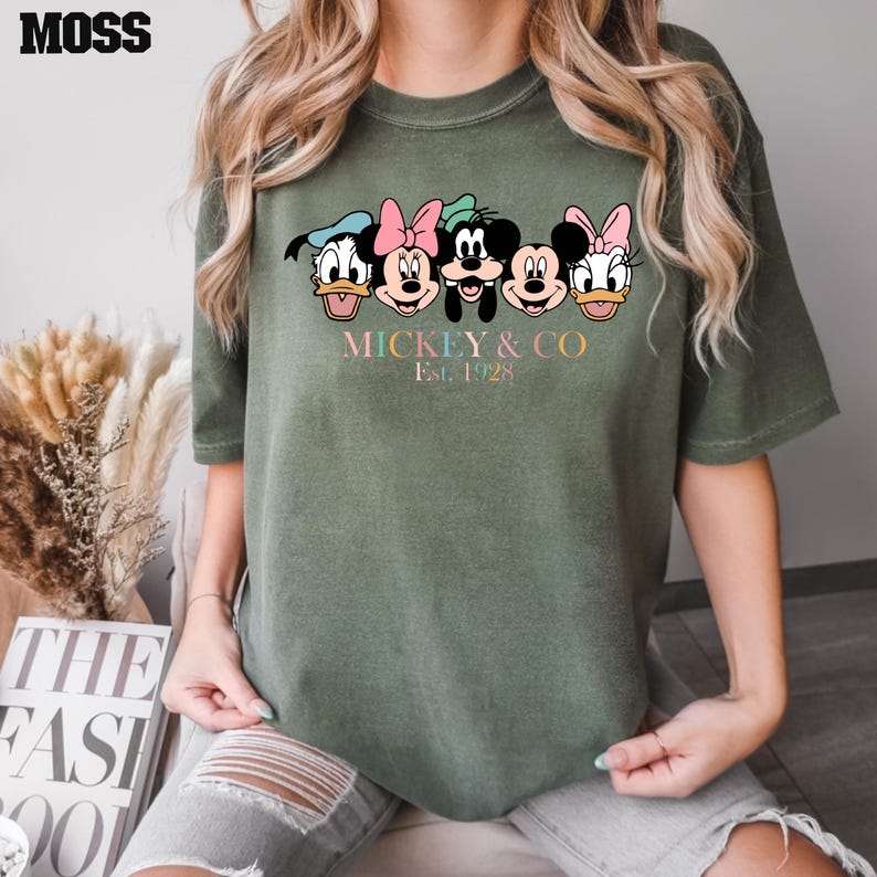 May include: Green t-shirt with a graphic of Mickey Mouse, Minnie Mouse, Donald Duck, Goofy, and Pluto. The text "Mickey & Co. Est. 1928" is printed below the graphic.