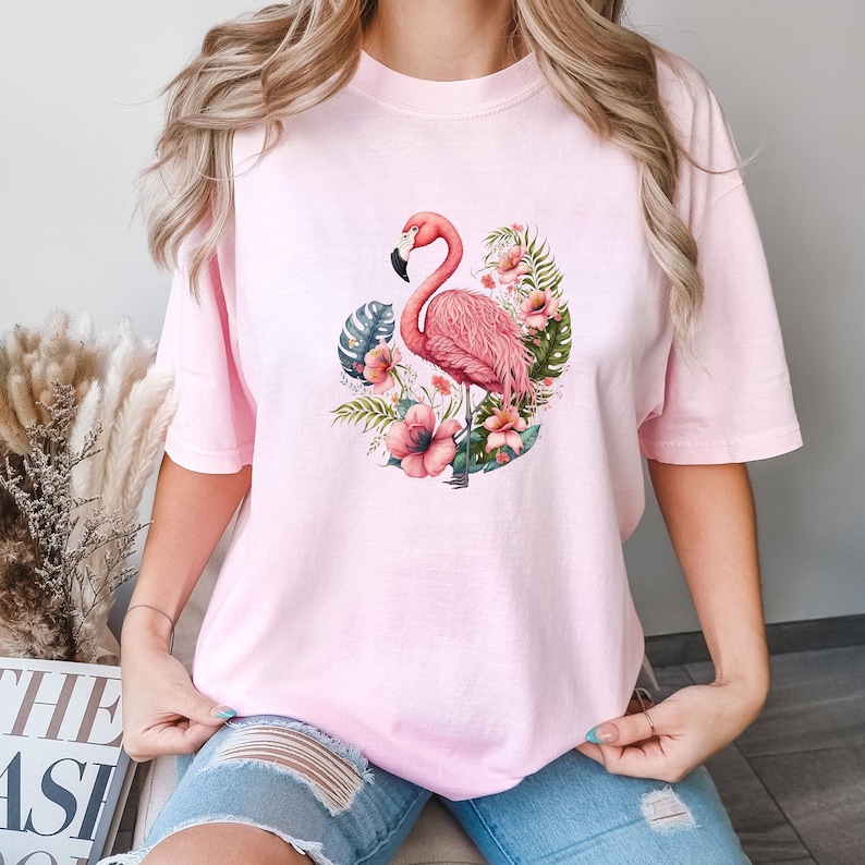K&ouml;nnte beinhalten: Rosa T-Shirt mit einem Flamingo- und tropischen Blumen-Grafikdruck. Der Flamingo ist rosa und steht auf einem Bein. Die Blumen sind rosa, gelb und wei&szlig;.