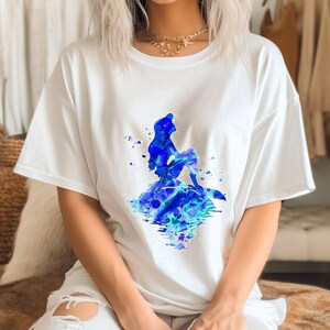 Puede incluir: Camiseta blanca con una ilustraci&oacute;n de acuarela azul de una sirena sentada en una roca con un fondo de agua azul y verde.