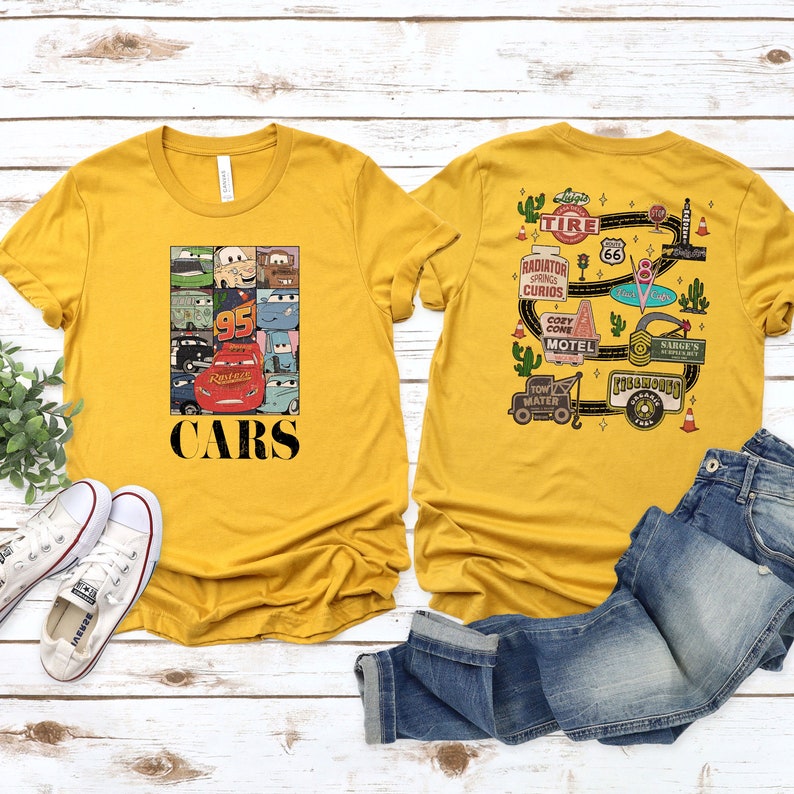 Puede incluir: Dos camisetas amarillas con tem&aacute;tica de Cars. La camiseta delantera tiene un collage de personajes de Cars y el texto "CARS". La camiseta trasera tiene un mapa de Radiador Springs con el texto "TIRE", "RADIATOR SPRINGS CURIOS", "COZY CONE MOTEL", "SARGE'S SPEED SHOP" y "TOW MATER".