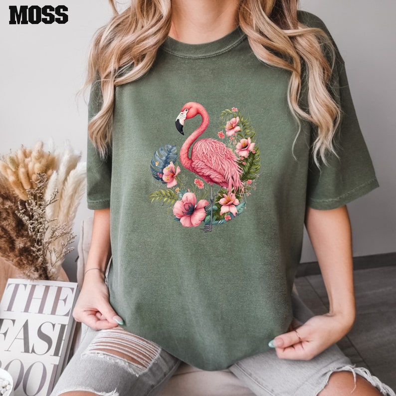 K&ouml;nnte beinhalten: Ein gr&uuml;nes T-Shirt mit einem pinkfarbenen Flamingo und einem Blumendesign. Der Flamingo steht auf einem Bein und hat den Kopf zur Seite gedreht. Die Blumen sind pink, wei&szlig; und gelb.