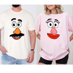 May include: Two t-shirts, one cream-colored and one pink, each featuring a cartoon face design. The cream shirt has a black mustache, orange nose, and red mouth. The pink shirt has eyelashes, a pink nose, and a red mouth.