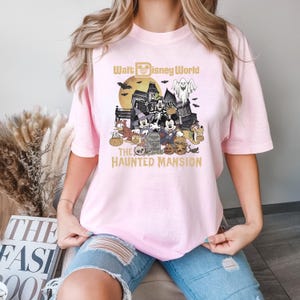 Puede incluir: Camiseta rosa con un gr&aacute;fico de Mickey Mouse y sus amigos frente a la Mansi&oacute;n Encantada en Walt Disney World. El texto "Walt Disney World" y "The Haunted Mansion" est&aacute; impreso en dorado.