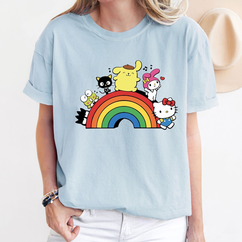 Pode incluir: Uma camiseta azul claro com um arco-&iacute;ris e personagens de desenhos animados. Os personagens incluem um cachorro amarelo, um gato preto, um coelho rosa, um sapo verde e um gato branco com um la&ccedil;o vermelho.