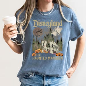 May include: A blue t-shirt with a vintage-style graphic of Mickey Mouse and a ghost in front of the Haunted Mansion at Disneyland. The text "Disneyland" and "The Haunted Mansion" are printed on the shirt.