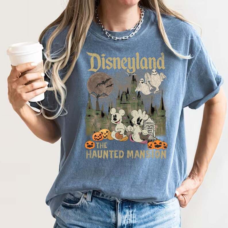 May include: A blue t-shirt with a vintage-style graphic of Mickey Mouse and a ghost in front of the Haunted Mansion at Disneyland. The text "Disneyland" and "The Haunted Mansion" are printed on the shirt.
