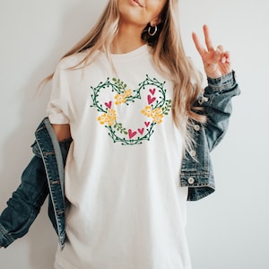 K&ouml;nnte beinhalten: Wei&szlig;es T-Shirt mit einem gr&uuml;nen Blumendesign in Form von Mickey-Maus-Ohren. Das Design enth&auml;lt gelbe Blumen und rosa Herzen.