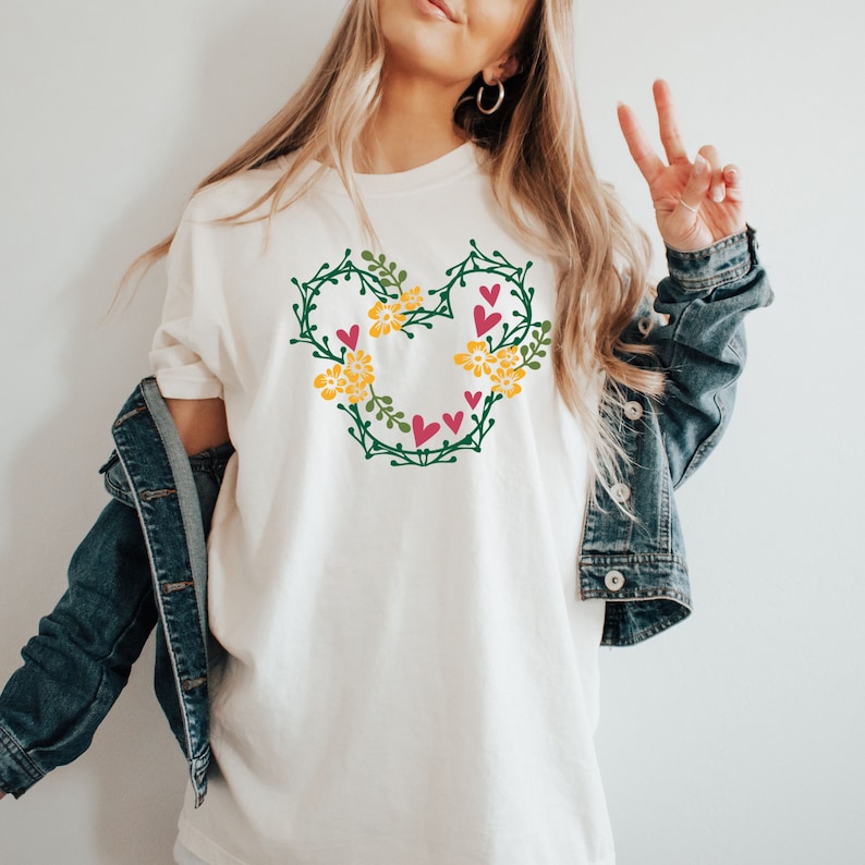 K&ouml;nnte beinhalten: Wei&szlig;es T-Shirt mit einem gr&uuml;nen Blumendesign in Form von Mickey-Maus-Ohren. Das Design enth&auml;lt gelbe Blumen und rosa Herzen.