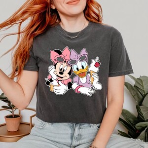Puede incluir: Una camiseta negra recortada con un estampado gr&aacute;fico de Minnie Mouse y Daisy Duck con gafas de sol y sosteniendo conos de helado.
