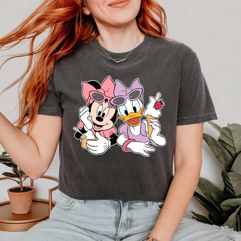 Puede incluir: Una camiseta negra recortada con un estampado gr&aacute;fico de Minnie Mouse y Daisy Duck con gafas de sol y sosteniendo conos de helado.