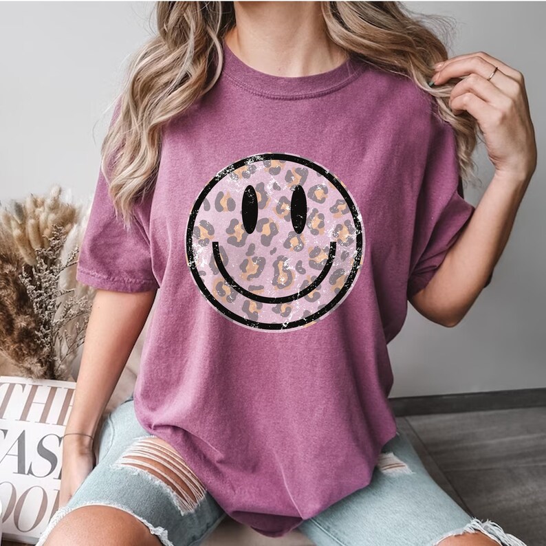 May include: A mauve t-shirt with a distressed, black-outlined smiley face graphic. The smiley face is filled with a pink and brown leopard print.