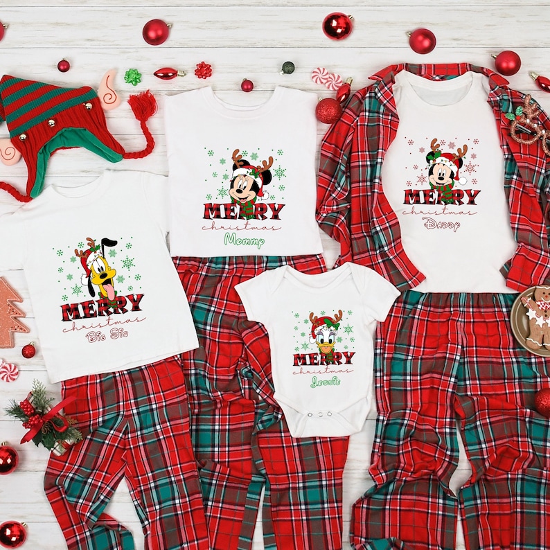Op de afbeelding: Een bijpassende kerstpyjamaset voor een gezin, bestaande uit rode en groene geruite broeken en witte shirts met een Mickey Mouse en Pluto ontwerp. De shirts hebben de tekst "Merry Christmas" en de namen "Mommy", "Daddy", "Big Sis" en "Little Sis".