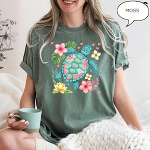 May include: Moss green t-shirt featuring a turquoise sea turtle with a floral design on its shell. The shirt is adorned with pink, yellow, and white flowers, green leaves, and small pink hearts. The word "MOSS" is in a speech bubble.