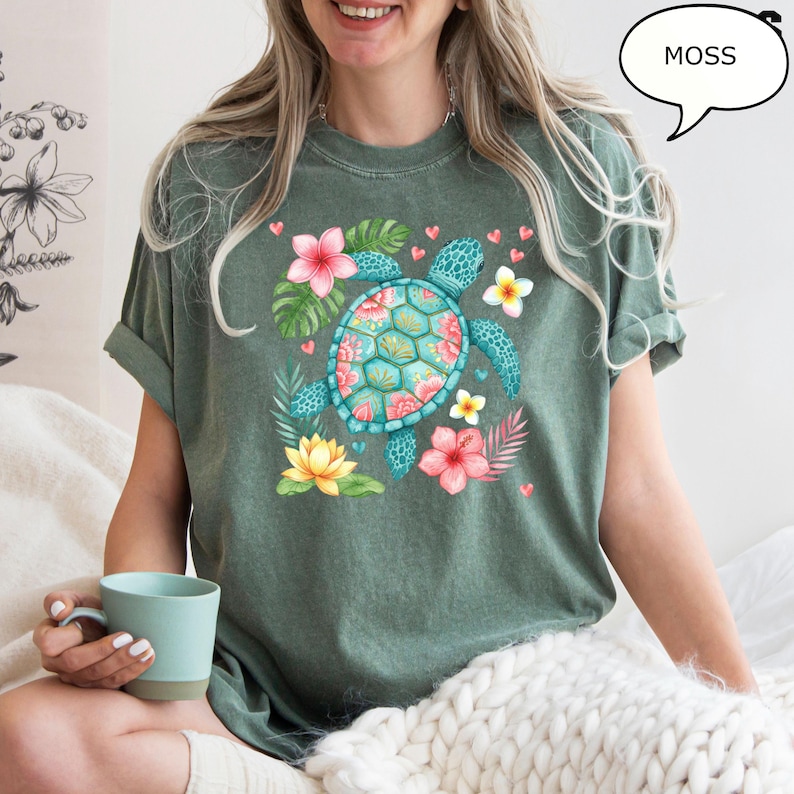 May include: Moss green t-shirt featuring a turquoise sea turtle with a floral design on its shell. The shirt is adorned with pink, yellow, and white flowers, green leaves, and small pink hearts. The word "MOSS" is in a speech bubble.