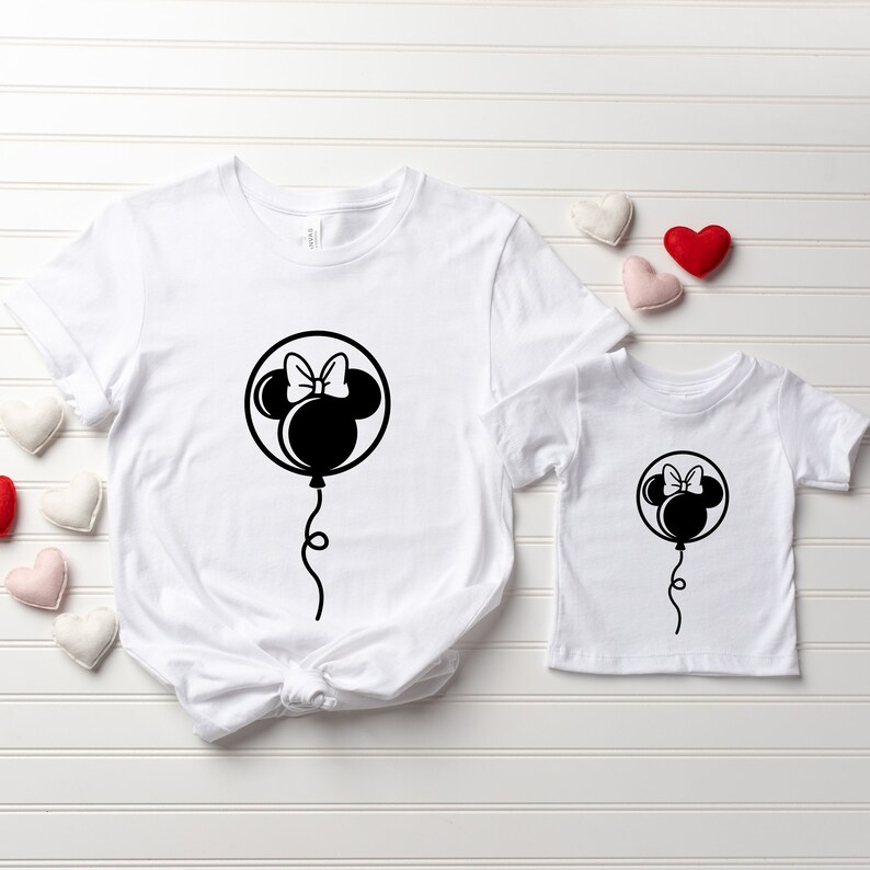 Pode incluir: Duas camisetas brancas com uma silhueta preta da cabe&ccedil;a do Mickey Mouse dentro de um bal&atilde;o. O bal&atilde;o tem um la&ccedil;o no topo. As camisetas est&atilde;o sobre um fundo de madeira branca.