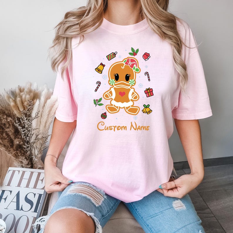May include: Pink t-shirt with a gingerbread person design. The gingerbread person is wearing a red heart and a green holly wreath. The shirt also has a variety of Christmas-themed decorations, including candy canes, bells, and gift boxes. The text "Custom Name" is printed below the gingerbread person.