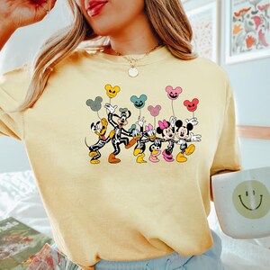 May include: A yellow t-shirt with a graphic print of Mickey Mouse and friends dressed as skeletons holding colorful balloons. The characters are smiling and appear to be celebrating Halloween.