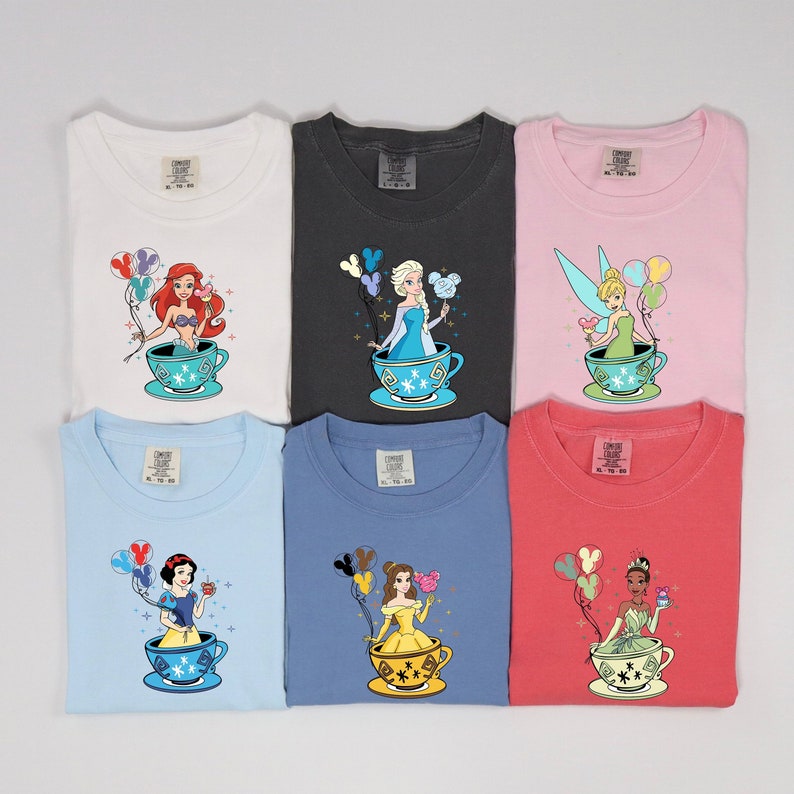 May include: Six different t-shirts featuring Disney princesses in teacups with balloons. The princesses are Ariel, Elsa, Tinkerbell, Snow White, Belle, and Tiana. The shirts are in various colors, including white, black, pink, light blue, blue, and red.