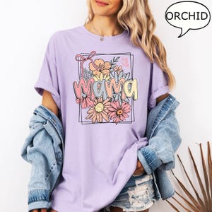 May include: A lavender t-shirt with a floral design and the word "mama" in a colorful font. The design features a bouquet of flowers with a pink bow.