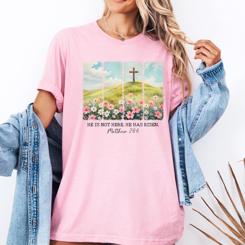 May include: Pink t-shirt featuring a graphic of a wooden cross on a hill with flowers. The text "He is not here. He has risen. Matthew 28:6" is printed below the graphic.