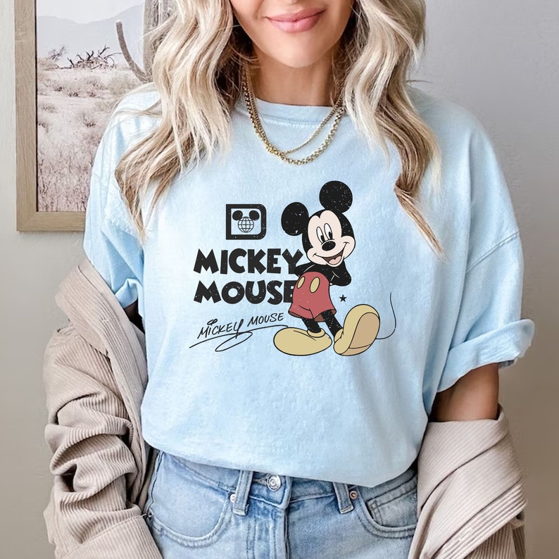May include: A light blue t-shirt with a vintage Mickey Mouse graphic. The graphic features Mickey Mouse in a red and yellow outfit with a black outline. The text "Mickey Mouse" is printed in black below the graphic. The shirt has a relaxed fit and short sleeves.